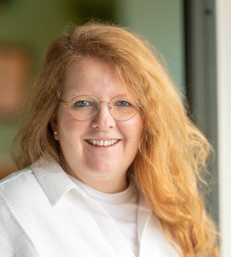eine lächelnde rothaarige Frau trägt eine weiße Bluse und eine Brille