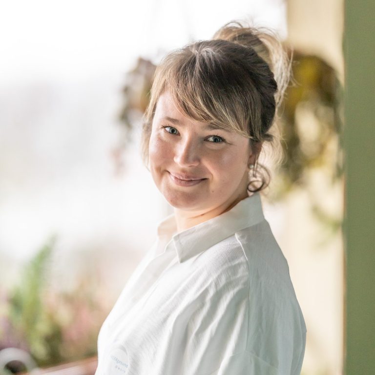 eine blonde Frau lächelt in die Kamera und trägt eine weiße Bluse